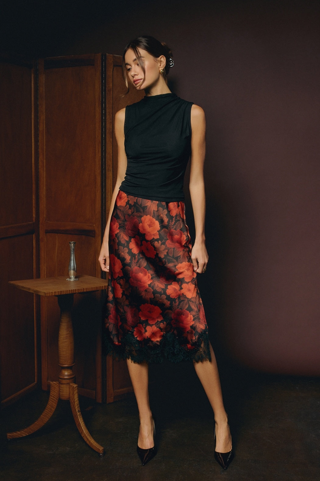 A model stands against a light gray studio background wearing a dark brown leather-look collared shirt and a leopard-print midi skirt with black lace trim and black strappy heels.