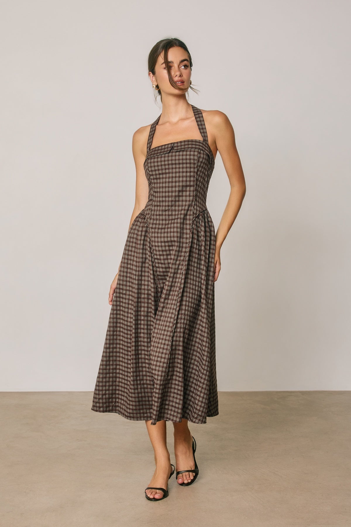 A woman modeling a brown and black checkered halter midi dress with a flared skirt, standing against a neutral light gray studio background.