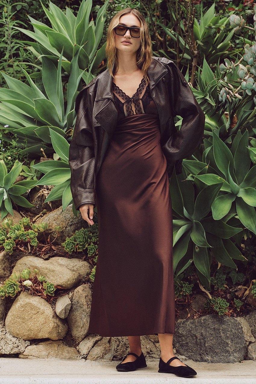 A woman stands outdoors in front of green succulents and rocks, wearing a dark brown satin midi dress with a lace-trimmed neckline, a black leather jacket, black flats, and sunglasses.