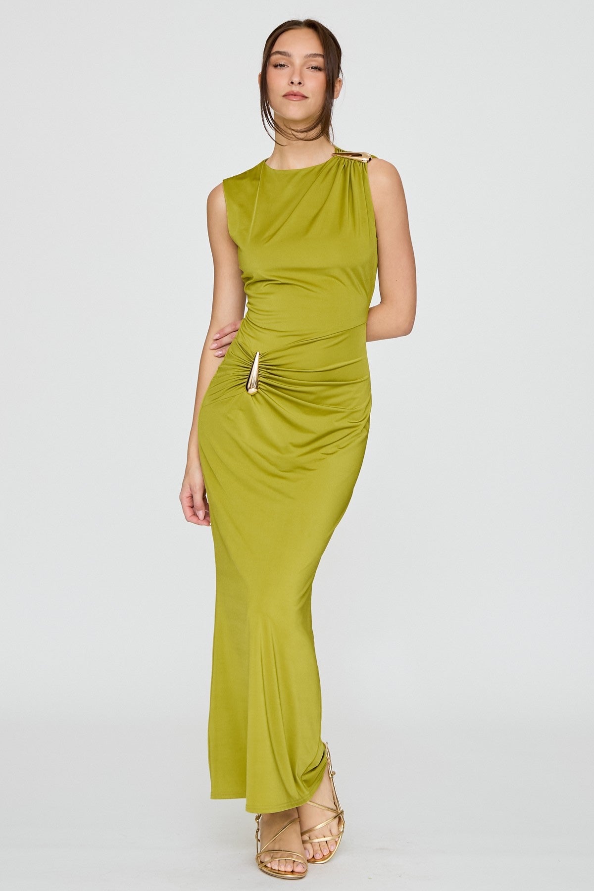 A woman modeling a sleeveless olive green maxi dress with a draped, gathered waist detail and a high neckline, paired with gold strappy heels, against a light gray studio background.