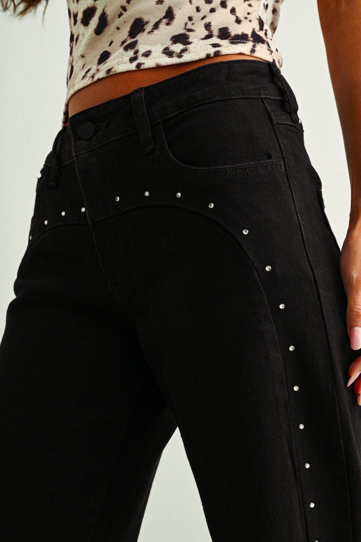 Close-up of a person wearing black jeans with silver stud embellishments along the front and side seams, paired with a leopard-print cropped top against a light gray background.