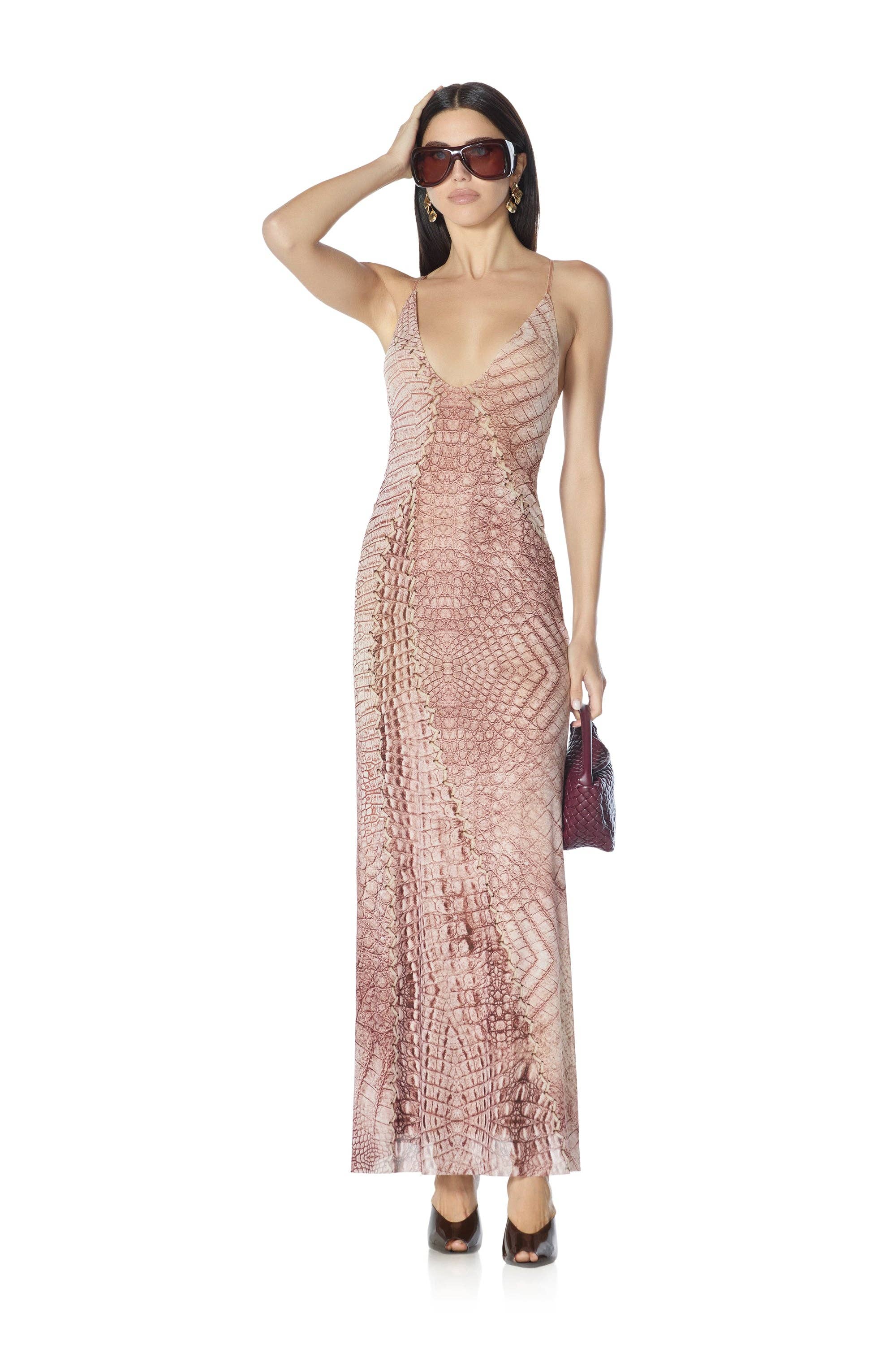 A woman modeling a floor-length, spaghetti-strap dress with a snakeskin pattern in dusty pink and brown tones, styled with dark sunglasses, a small dark handbag, and dark pointed shoes against a plain white background. AFRM BRAND
