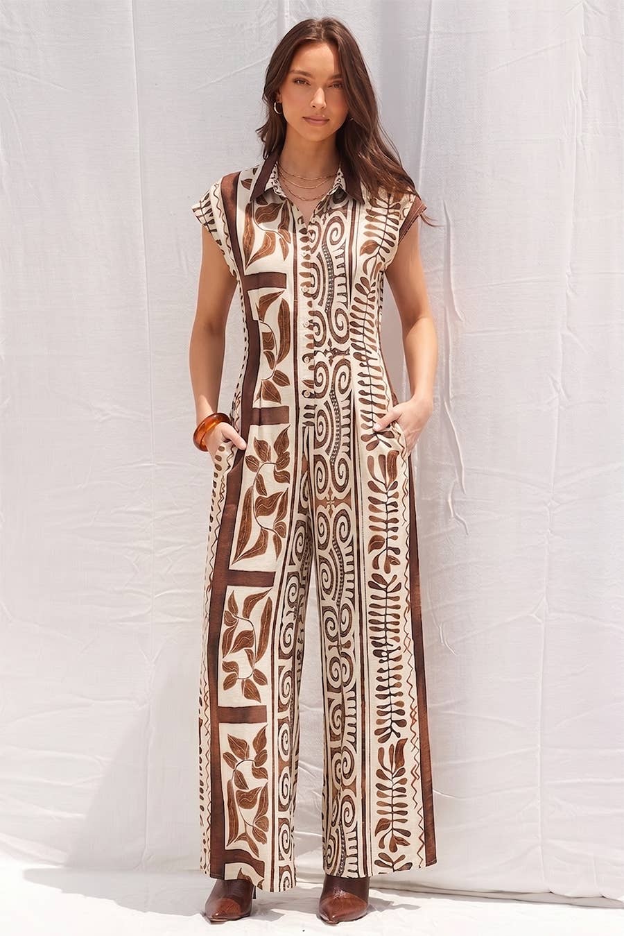 A woman modeling a sleeveless, wide-leg jumpsuit with a brown and beige tribal-inspired geometric pattern, standing against a white fabric backdrop.