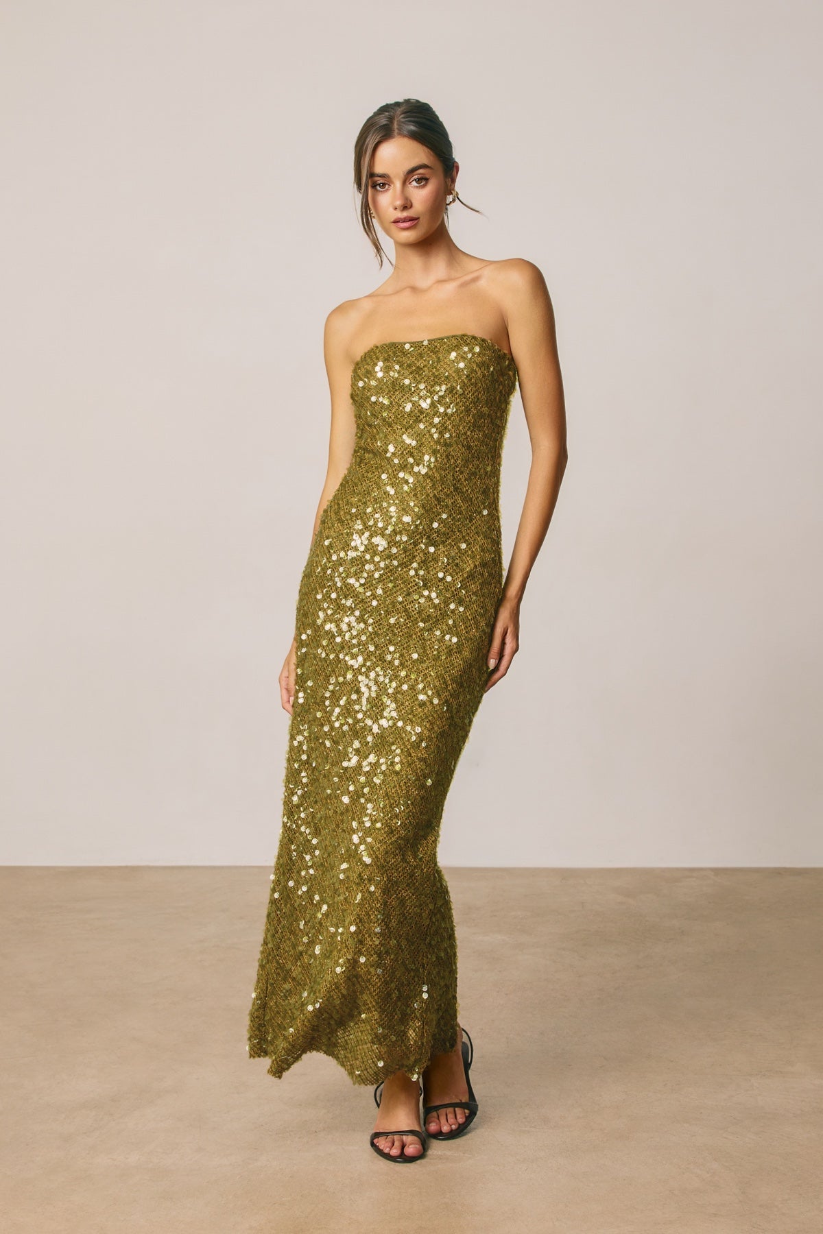 A woman modeling a strapless olive green sequined maxi dress with a fitted bodice and flared skirt, paired with black strappy heels, against a neutral beige studio backdrop.