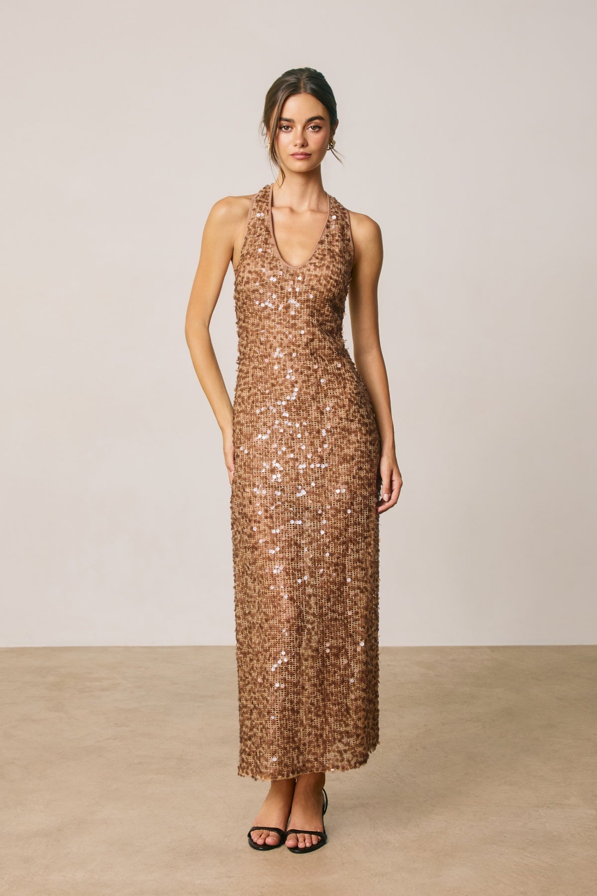 A woman modeling a sleeveless, halter-neck, floor-length sequin dress in a warm metallic bronze color, standing against a neutral beige studio backdrop.