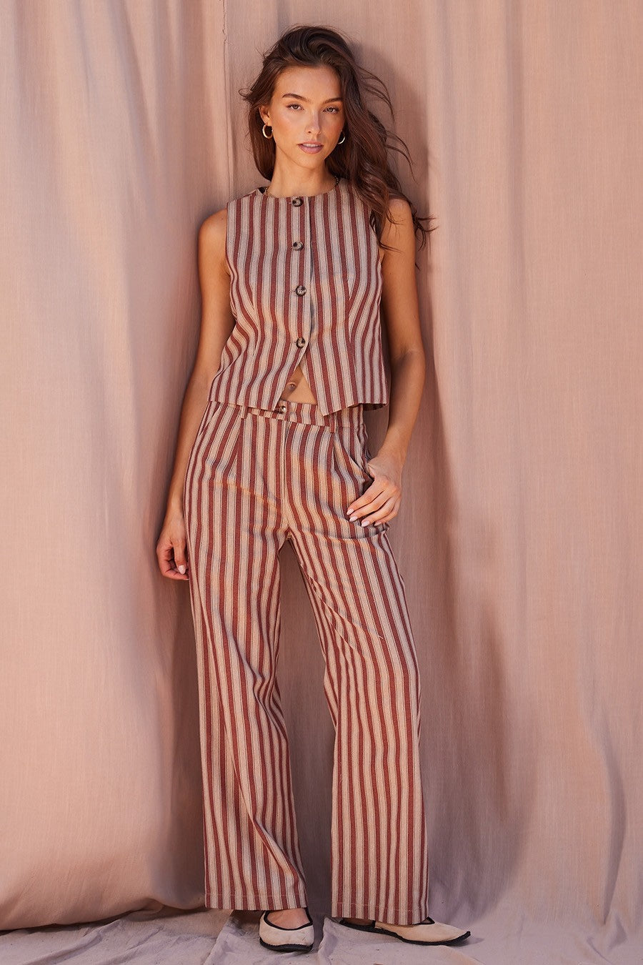 A woman modeling a sleeveless striped jumpsuit with a button-front bodice and wide-leg pants, posed against a draped dusty pink fabric backdrop.