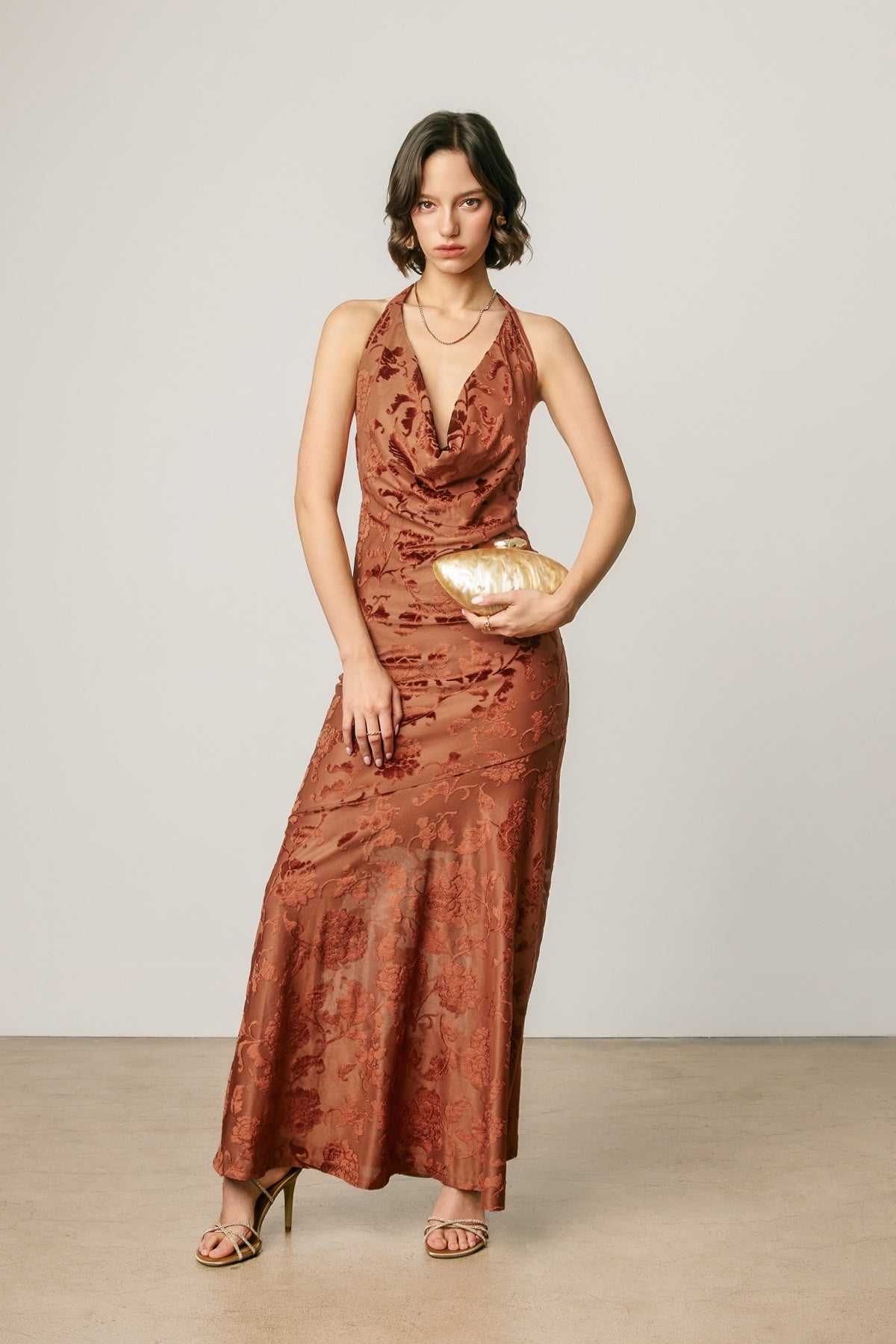 A woman modeling a rust-brown halter-neck maxi dress with a textured, tonal pattern, holding a gold clutch against a neutral studio backdrop.
