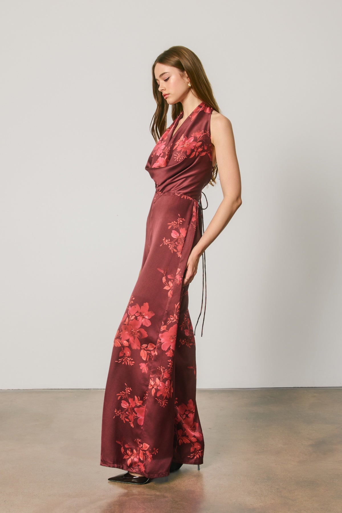 A woman modeling a sleeveless, floor-length wrap-style maxi dress in deep burgundy with a red floral print, standing in a studio with a light gray wall and concrete floor.