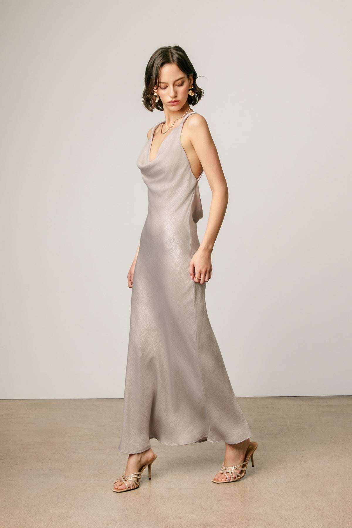 A woman modeling a floor-length satin slip dress in a champagne beige color, standing in a minimalist studio with a light gray wall and beige floor.