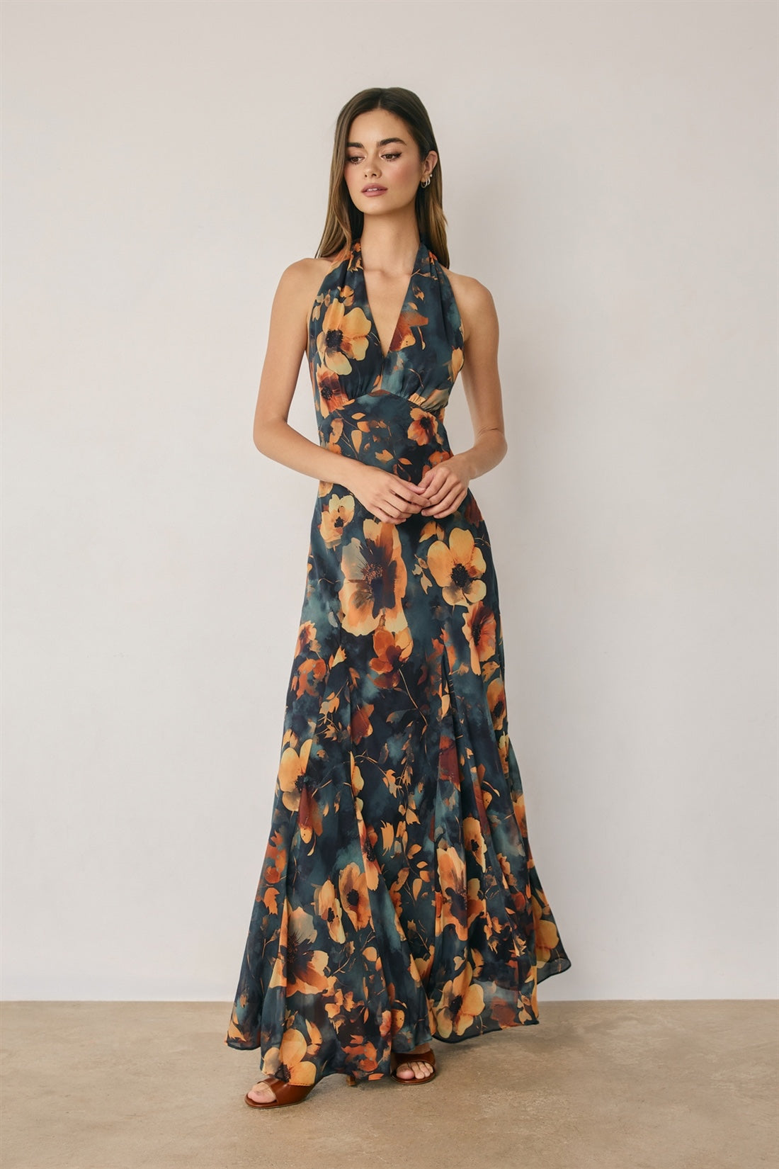 A woman modeling a sleeveless halter-neck maxi dress with a dark navy blue base and a large orange, yellow, and brown floral print, standing against a plain light beige wall.