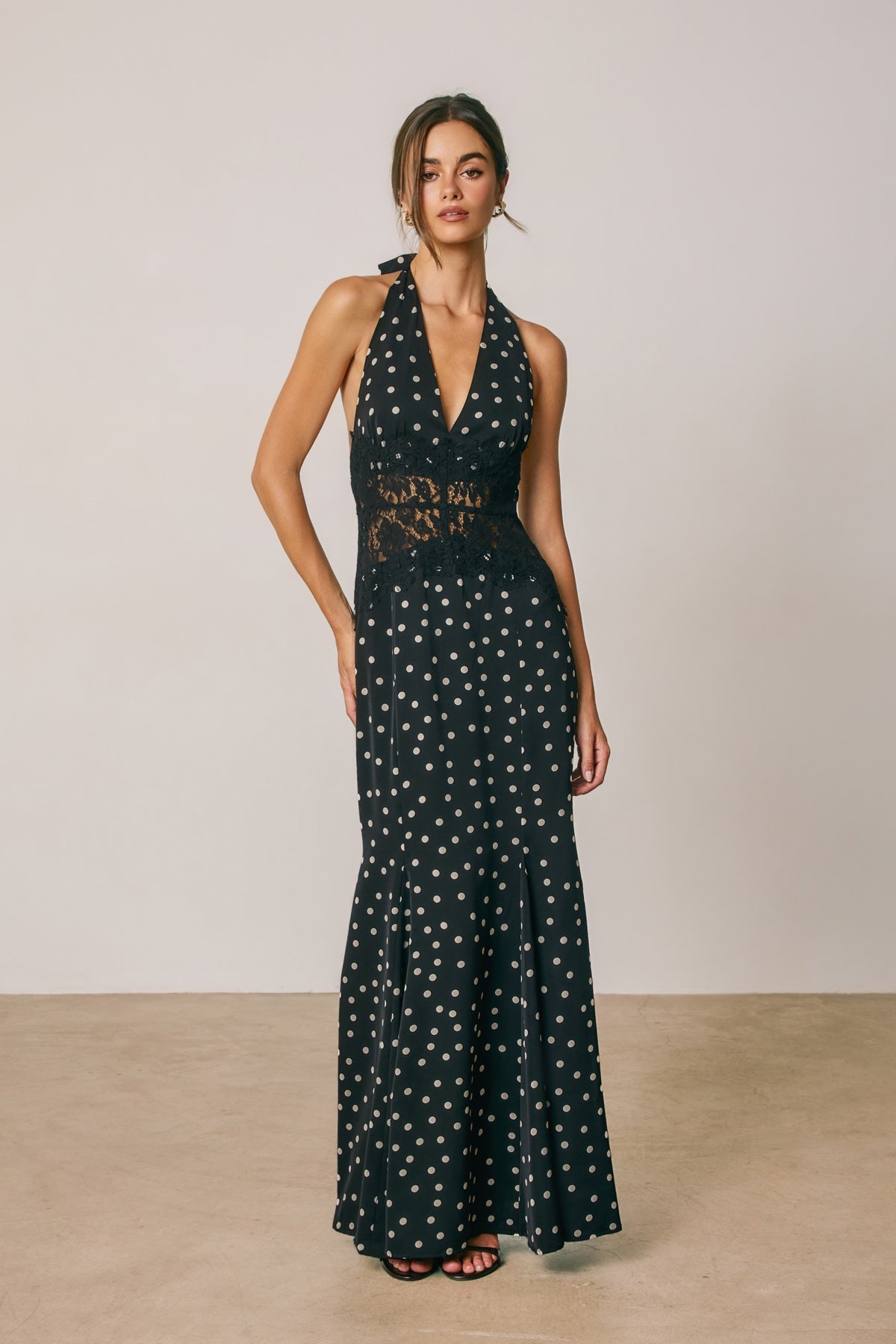 A woman modeling a sleeveless halter-neck maxi dress with a dark green base and white polka dots, featuring a sheer lace waist panel, set against a neutral studio backdrop.