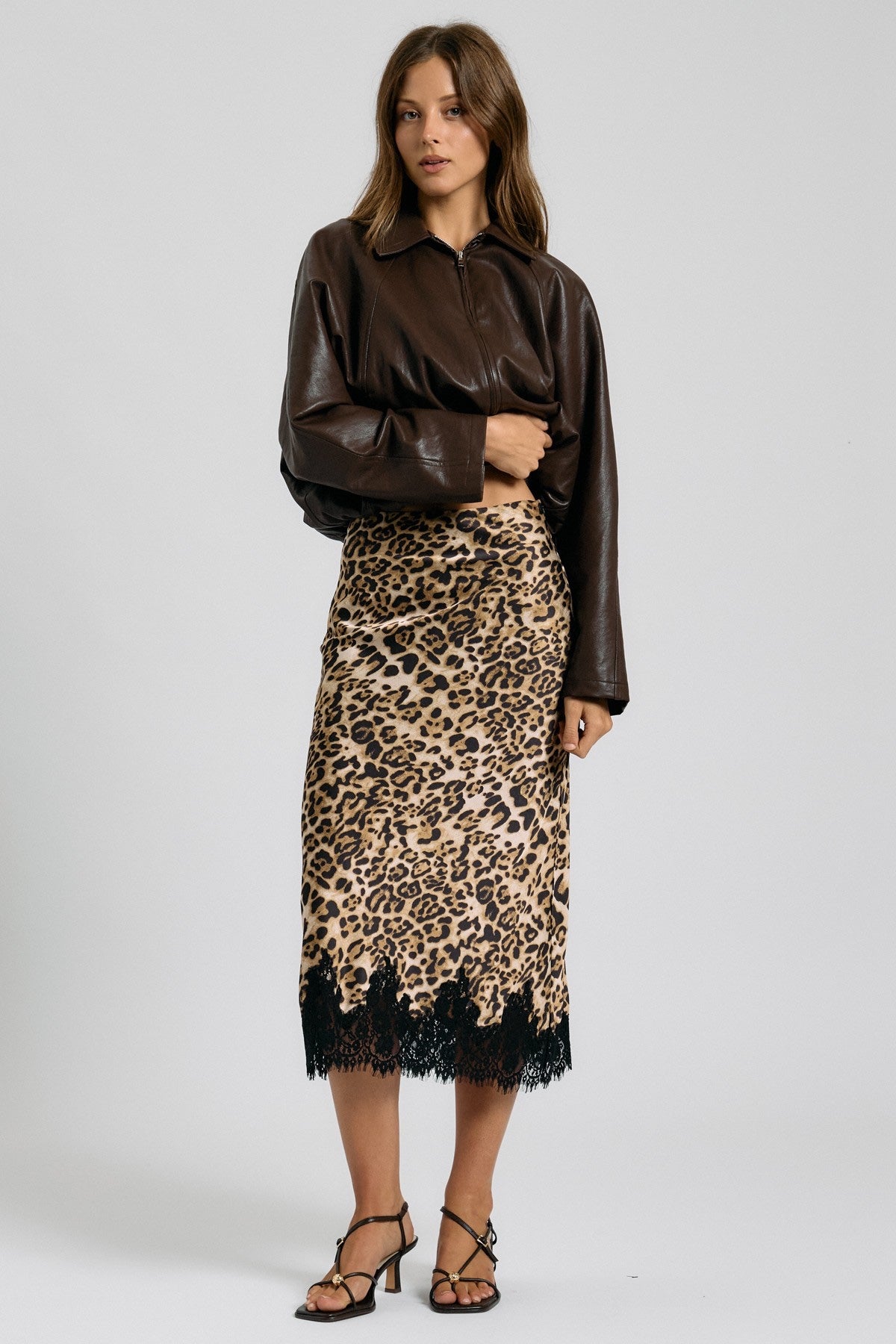A model stands against a light gray studio background wearing a dark brown leather-look collared shirt and a leopard-print midi skirt with black lace trim and black strappy heels.
