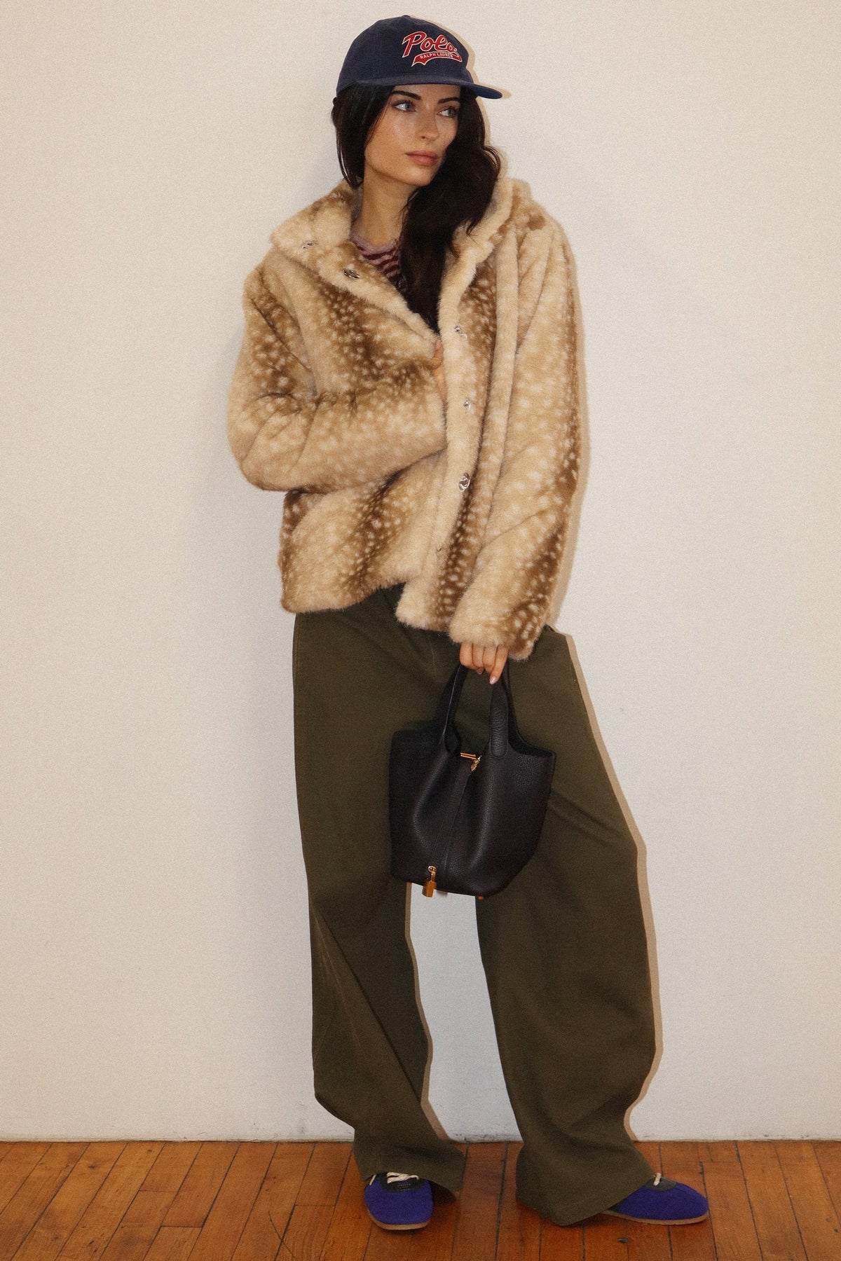 A woman stands against a plain off-white wall wearing a beige and brown mottled faux fur jacket, olive green wide-leg pants, a navy baseball cap, and blue shoes, while holding a black handbag.