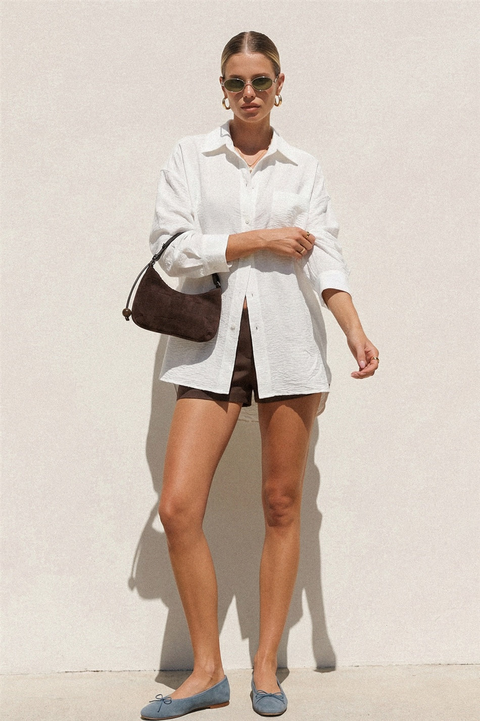 Woman wearing a white shirt, brown shorts, and blue flats against a light background