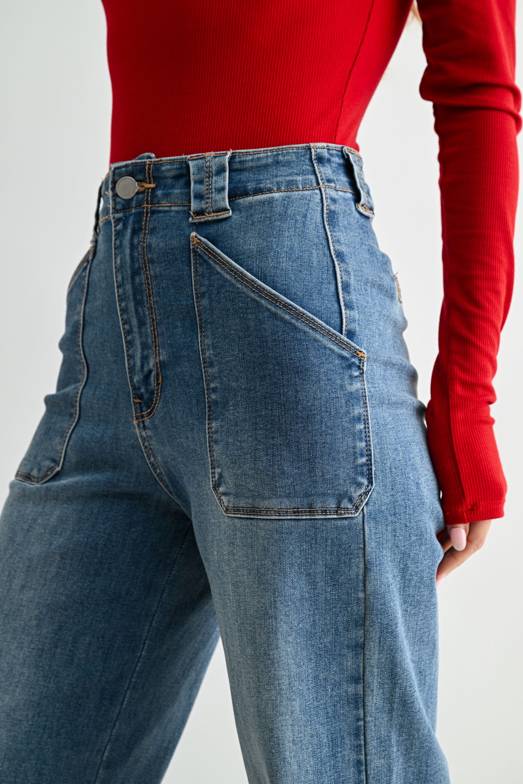 Close-up of a person wearing high-waisted blue jeans with a red ribbed long-sleeve top, showing the back pocket and waistband details against a light gray studio background.