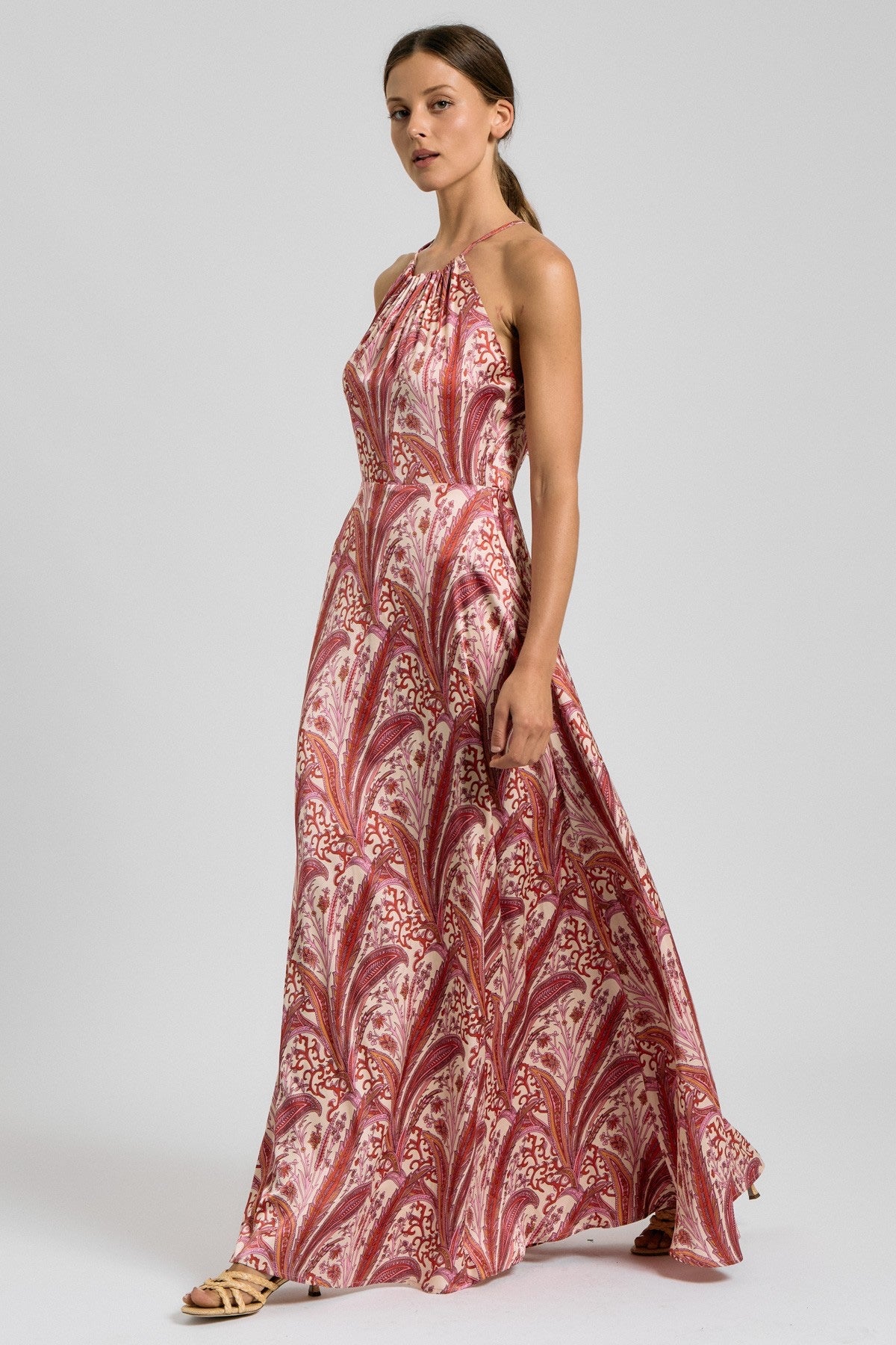 A woman modeling a sleeveless halter-neck maxi dress with a red, pink, and beige paisley pattern, standing against a light gray studio background.
