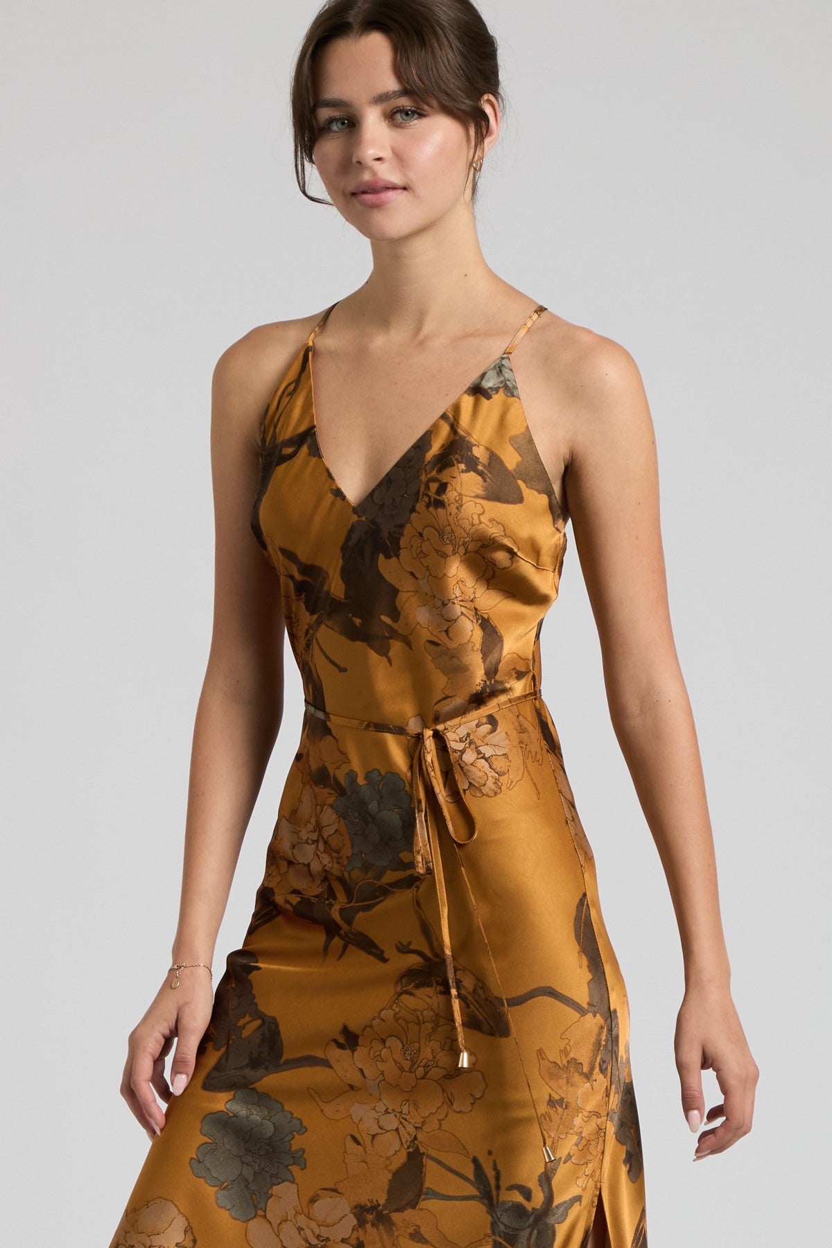 A woman modeling a sleeveless wrap-style midi dress with a tie waist, featuring a golden-brown satin-like fabric with a dark floral print, against a light gray studio background.