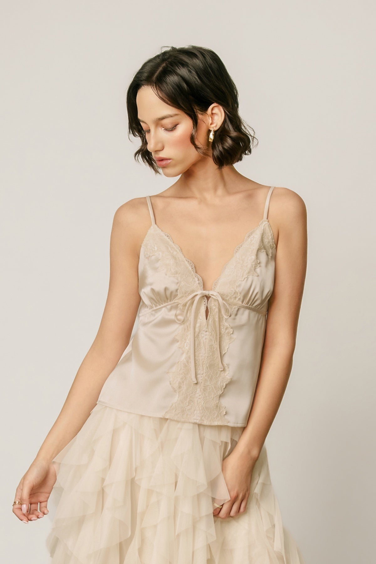 A woman wearing a light beige camisole top with lace detailing and a matching tiered tulle skirt, photographed against a plain light gray studio background.