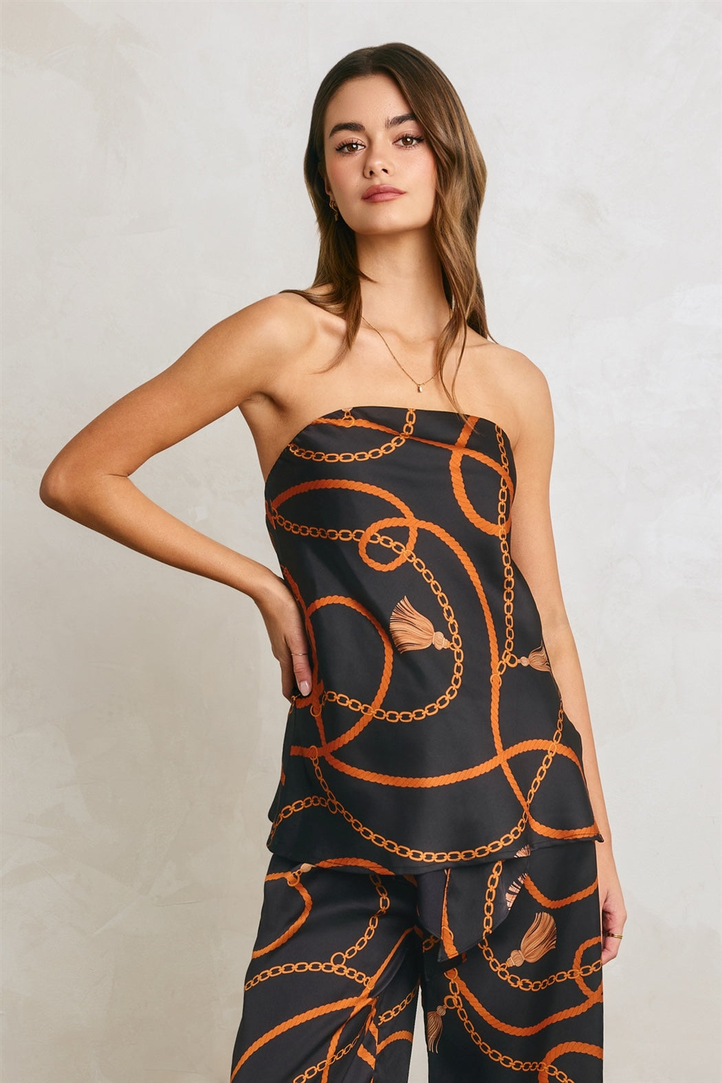 A woman modeling a strapless black jumpsuit with a bold orange chain-link pattern and small butterfly motifs, set against a neutral textured light gray background.