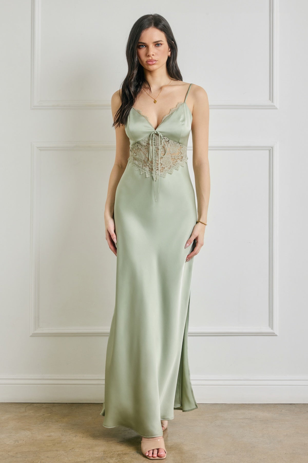 Woman wearing a long green dress with lace details against a white paneled wall.