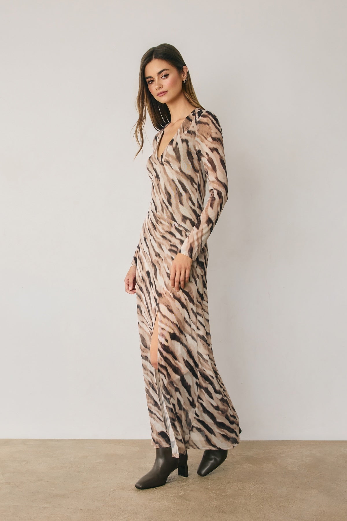 A woman models a long-sleeve maxi dress with a beige, brown, and black animal print pattern, standing against a neutral light gray wall and beige floor.